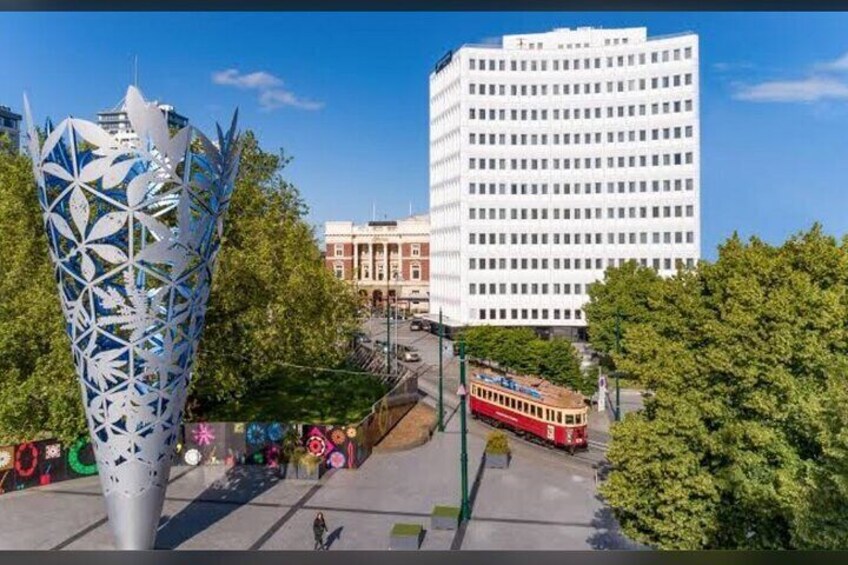 Eine moderne Skulptur, eine Straßenbahn und ein historisches Gebäude in einer Stadtansicht. 1
