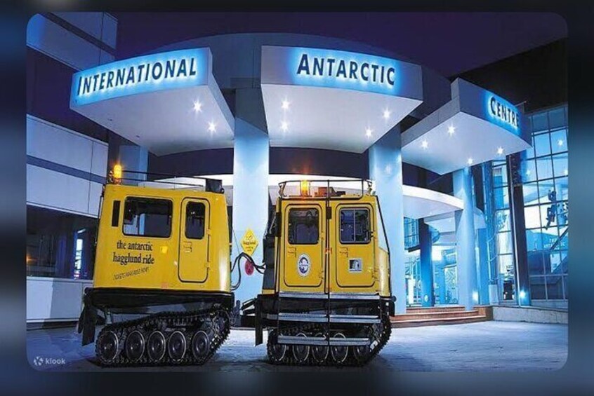 Zwei gelbe Fahrzeuge mit Ketten an einer Tankstelle, Schilder mit den Aufschriften 'International' und 'Antarktis'. 3
