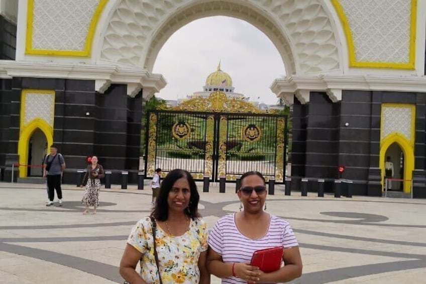 Two women standing in front of a large ornate gate with a golden emblem, with a domed building in the background. 2