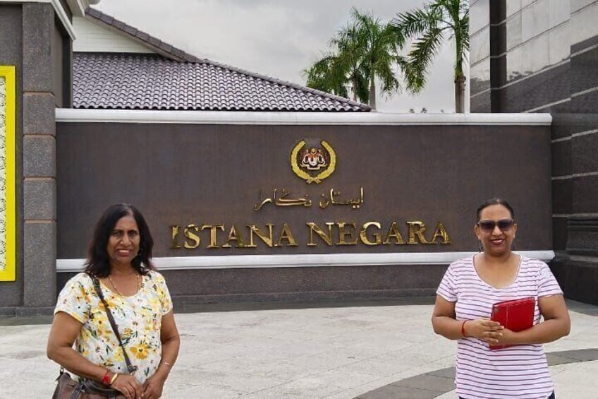 Two women standing in front of a sign with the text "ISTANA NEGARA" and a crest. 4