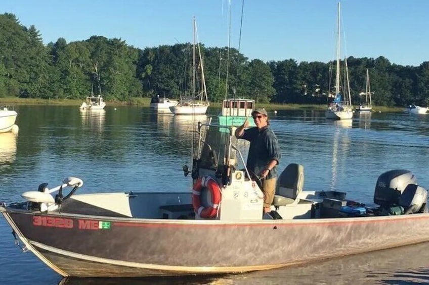 A man on a small boat with a motor, docked near a calm lake with several sailboats in the background. 3