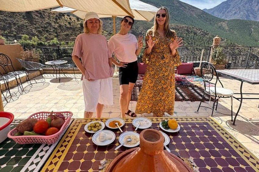 Trois personnes sont debout autour d’une table, devant de la nourriture et un grand pot en argile, avec des montagnes en arrière-plan. 3
