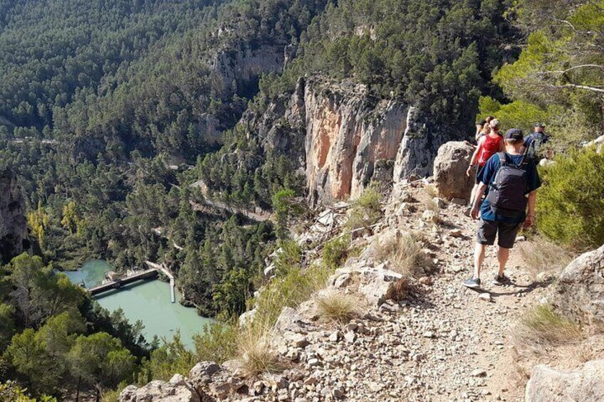 Un sendero rocoso para excursionismo con vista a un cañón, un pequeño embalse y un grupo de excursionistas. 1