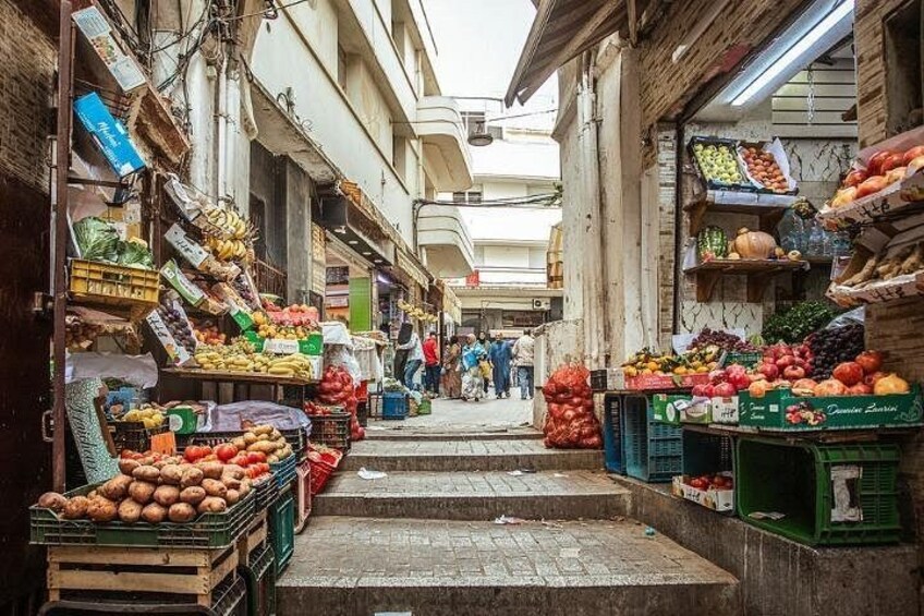 Un vicolo stretto fiancheggiato da bancarelle di frutta, con scalini che conducono a un livello più alto e edifici su entrambi i lati. 4