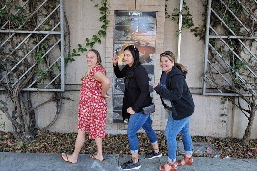 Three people walking on a sidewalk with a building and a poster in the background. 4
