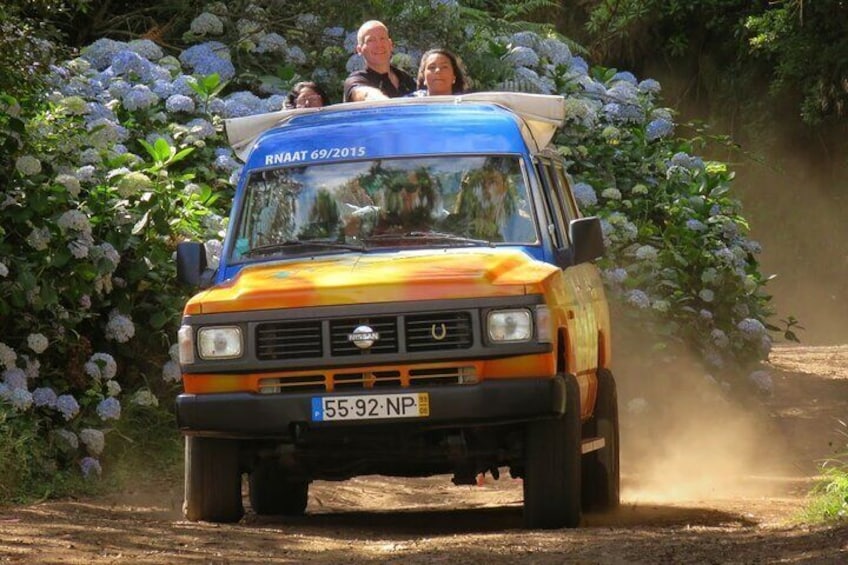 A vintage orange and blue Nissan pickup truck driving on a dirt road, surrounded by lush greenery and white flowers. 1