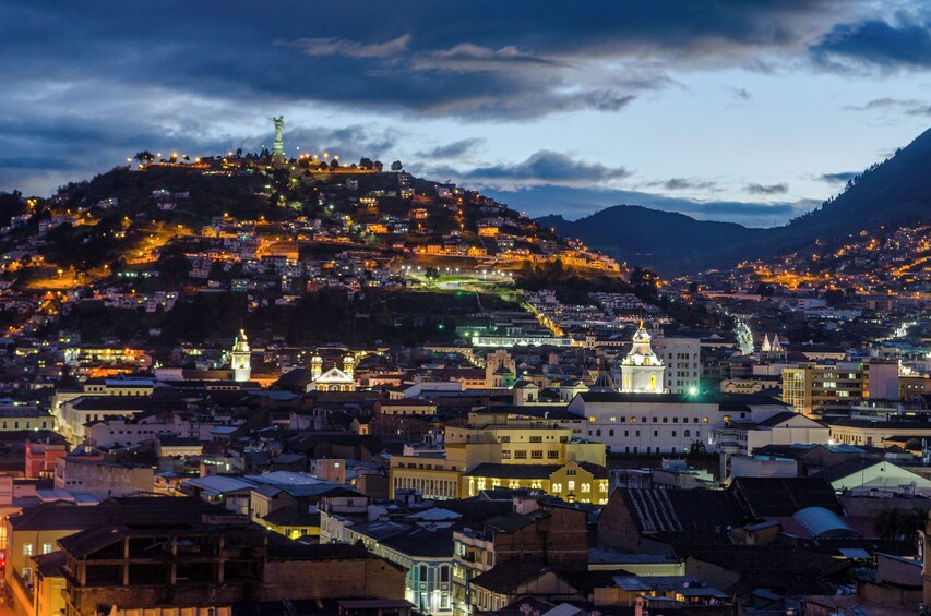 A cityscape at dusk with illuminated buildings, a prominent hilltop statue, and a cathedral. 4