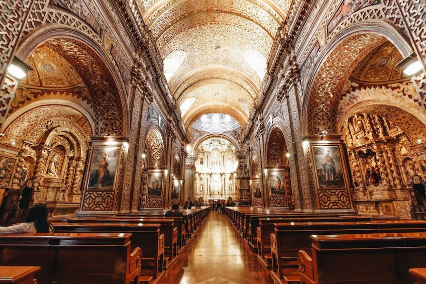 An ornate church interior with intricate gold detailing, wooden pews, and a central aisle leading to a high altar. 2