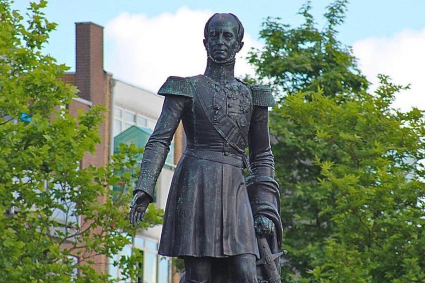 Une statue représentant une personne en uniforme militaire, portant une jupe, et se tenant à l’extérieur, avec des arbres et un bâtiment en arrière-plan. 3