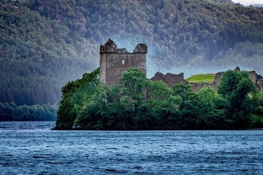 A historic stone castle surrounded by lush greenery and a forested hill in the background. 2