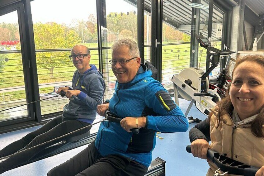 Tre personer træner på spinningcykler i et fitnesscenter med store vinduer med udsigt over et grønt område. 1