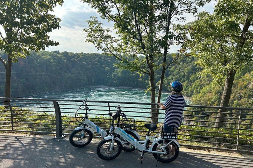 Una persona en una bicicleta estática con vista a un río, con árboles y una barandilla al frente. 5