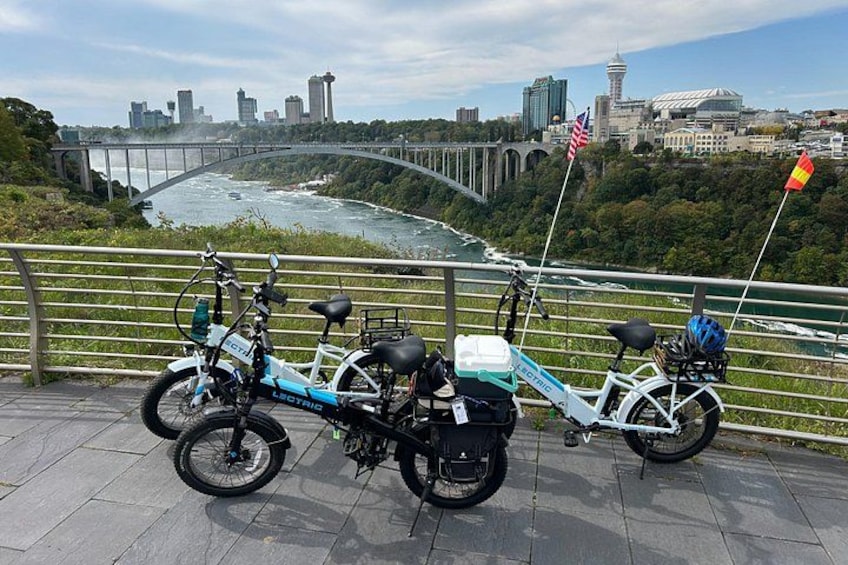 Dos bicicletas eléctricas estacionadas en un área pavimentada, con vista panorámica a un río, un puente y el horizonte de la ciudad. 3