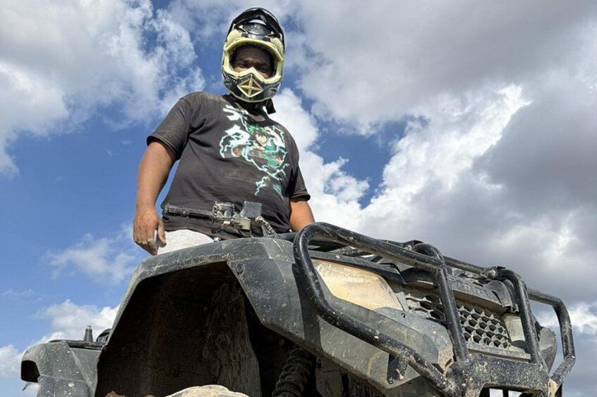 A person on an ATV with a helmet and a graphic t-shirt, standing on a vehicle with a cloudy sky in the background. 2