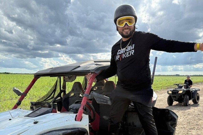 A person in a black shirt with 'RANGER' on it, standing next to a vehicle with a red frame, in a field with another vehicle in the background. 5
