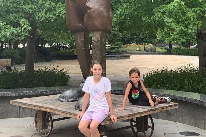 Deux enfants jouent sur une balançoire dans un parc, avec une grande statue de bronze en arrière-plan. 2