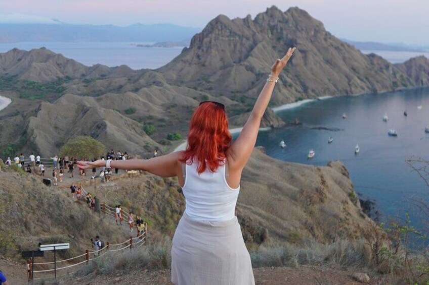 Eine rothaarige Person steht auf einer Klippe und blickt auf eine Küstenlandschaft mit Booten und felsigem Gelände. 1