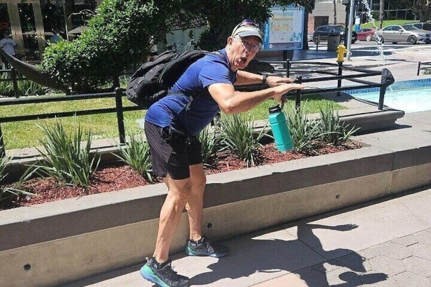 A person performing a stretching exercise outdoors, holding a water bottle, with a pool and greenery in the background. 5
