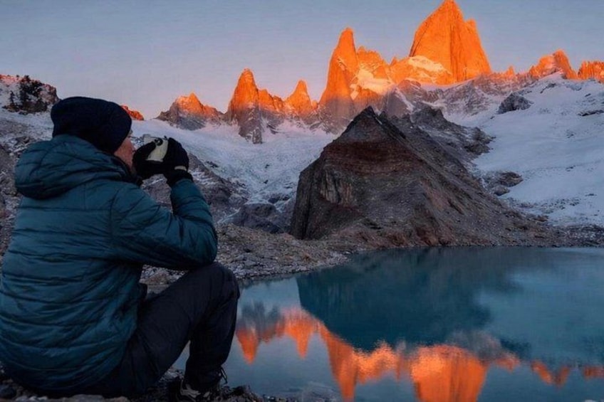 A person taking a photograph of a mountain range with sharp peaks and a reflective lake. 1