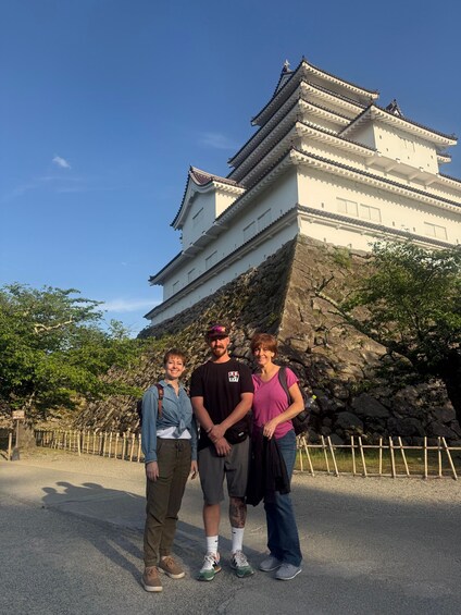 Three people standing in front of a white castle with multiple tiers and a steep rock face. 1