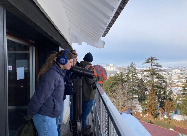 A person using a telescope on a balcony overlooking a cityscape with snow-covered trees. 4