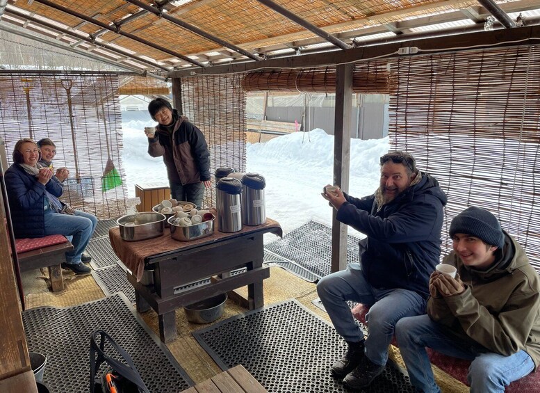 A group of people sitting and standing inside a covered structure with a table holding various containers and a stove. 3