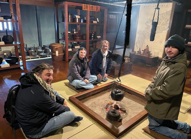 A group of people sitting around a traditional Japanese hearth with various cooking utensils in the background. 1