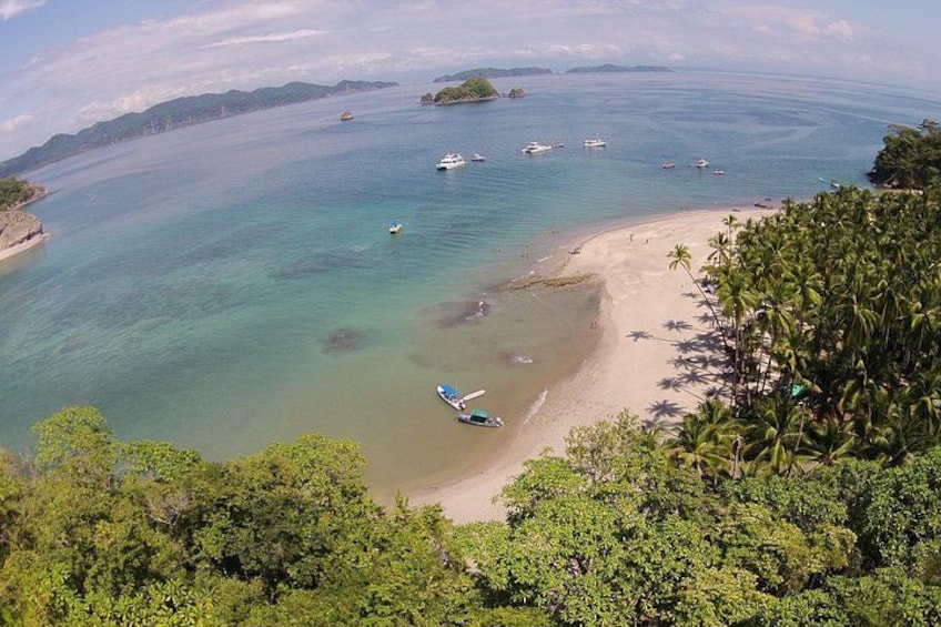 A coastal landscape with a beach, clear turquoise waters, boats, and dense tropical vegetation. 2