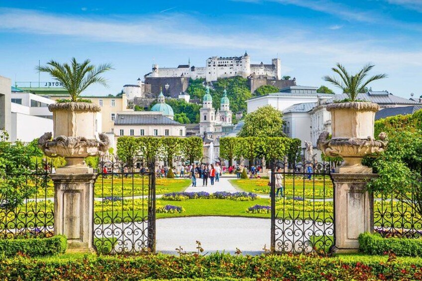 A historic castle on a hill, a well-maintained garden with a fountain, and a clear blue sky. 2