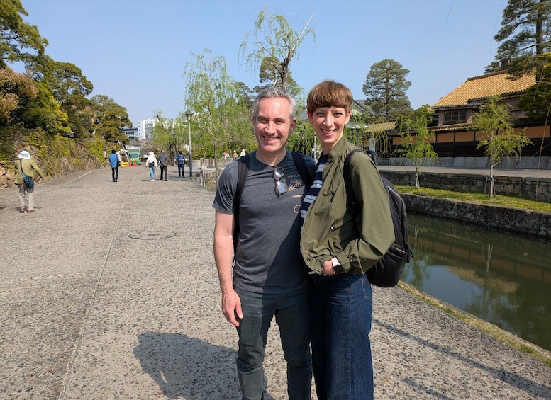 Two people standing by a water canal with trees and buildings in the background. 2