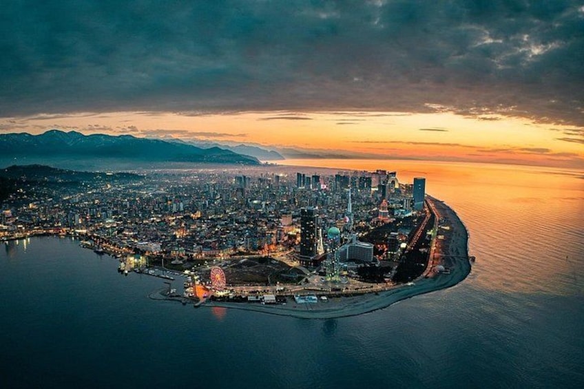 A coastal cityscape at sunset with a prominent Ferris wheel, a large body of water, and a mountain range in the background. 2