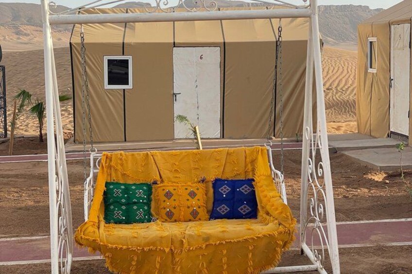 A yellow swing with patterned cushions in a desert setting with tents in the background. 2