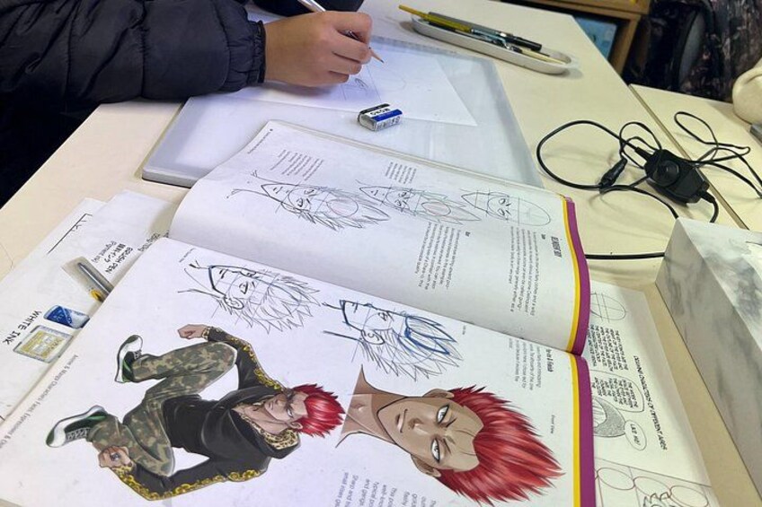 En person som ritar i ett skissblock med hjälp av olika verktyg, och en uppslagsbok ligger på ett skrivbord bredvid. 2