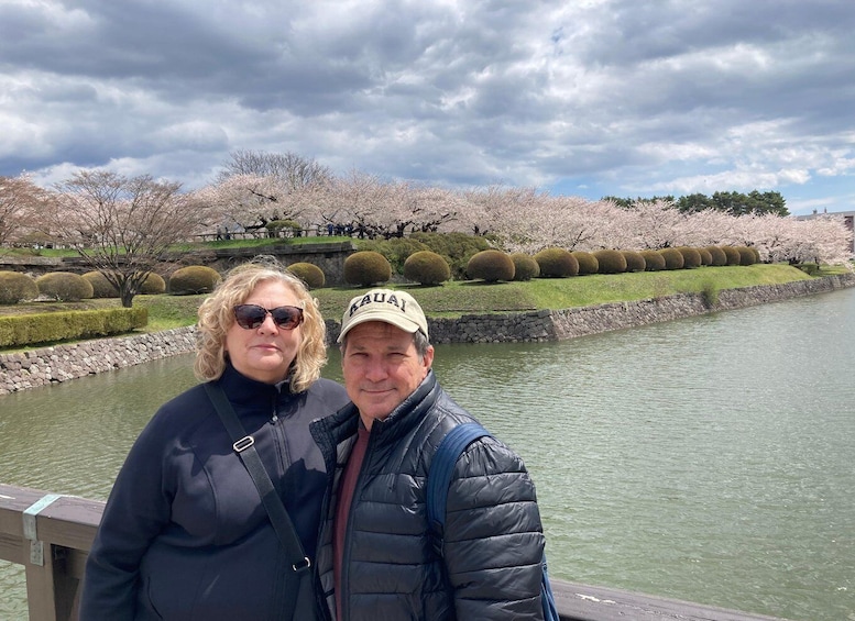 Two people standing by a river with cherry blossoms and a stone wall in the background. 4