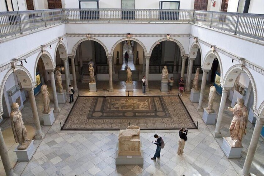 A museum courtyard with statues, a mosaic floor, and a balcony. 1