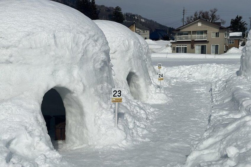 Schneebedeckte Iglus mit Nummern, ein verschneiter Weg und Häuser im Hintergrund. 2