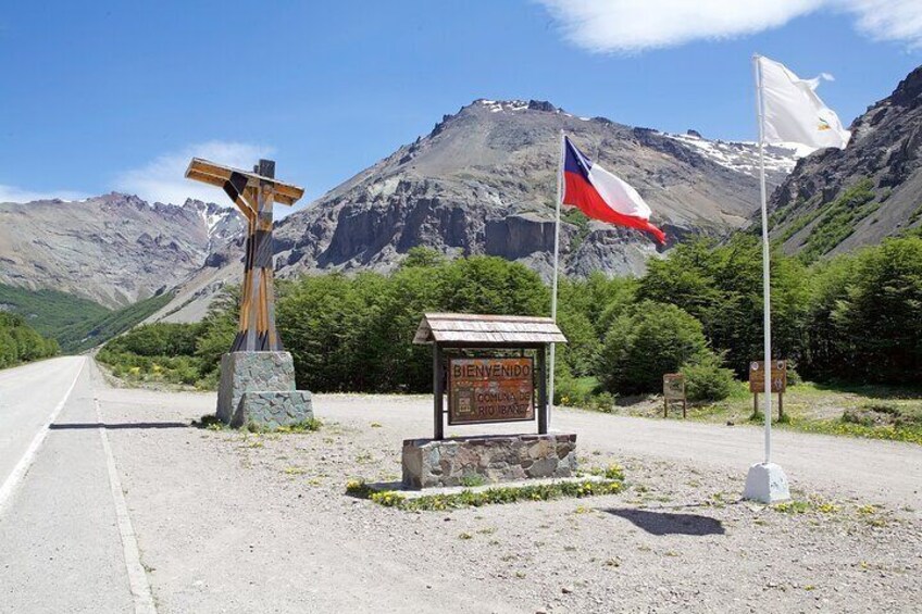 A cross, a sign, and flags on a roadside with mountains in the background. 2