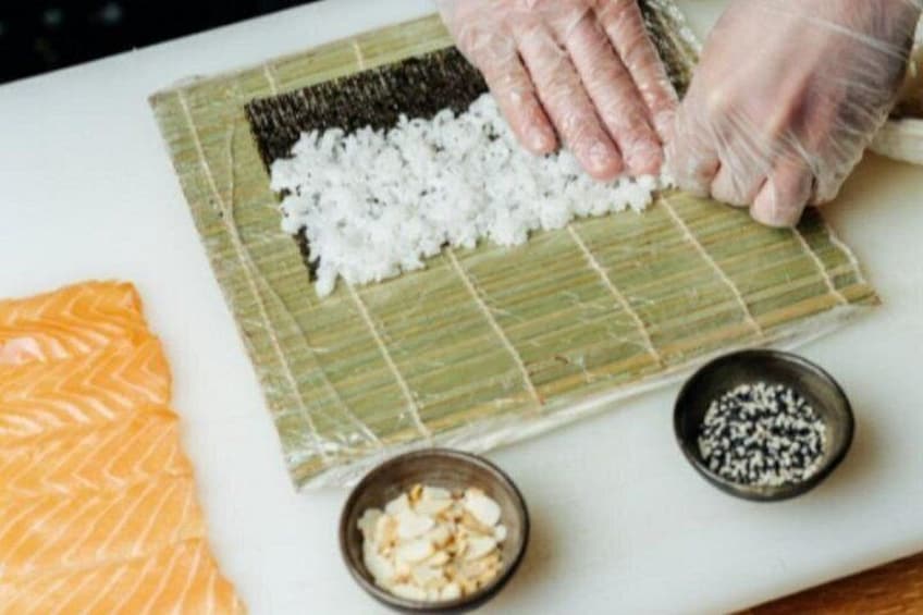 Hands preparing sushi with rice and seaweed on a cutting board, with salmon and two small bowls of condiments. 1