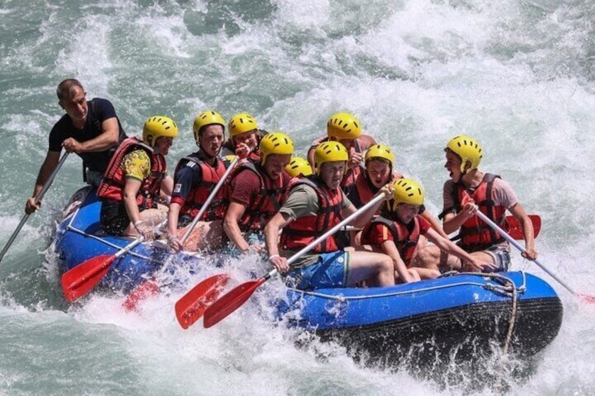 Eine Gruppe von Menschen in einem blauen Floß mit roten Paddeln, mit Helmen und Schwimmwesten, bewegt sich durch unruhiges Wasser. 1