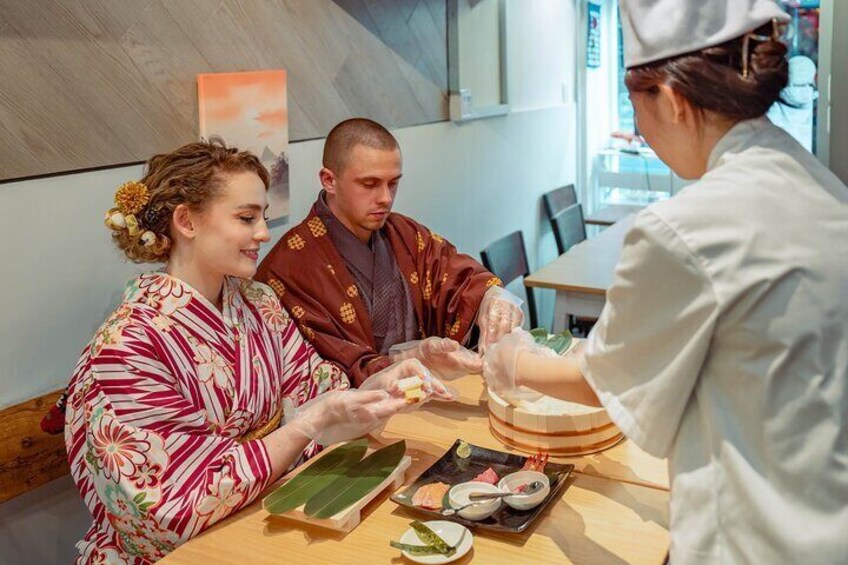 A person in a white uniform serving food to two customers at a table with sushi and a green leaf. 1