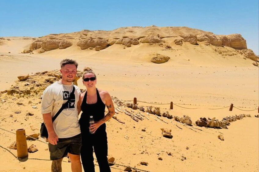 Two people standing in a desert with sand dunes and a fenced area in the background. 5