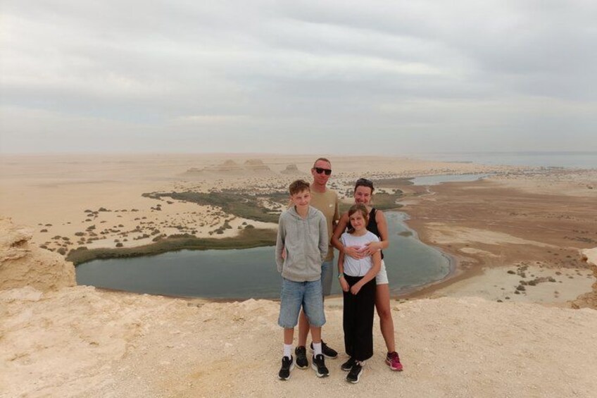 A family of four standing on a sandy terrain with a large body of water and distant hills. 3