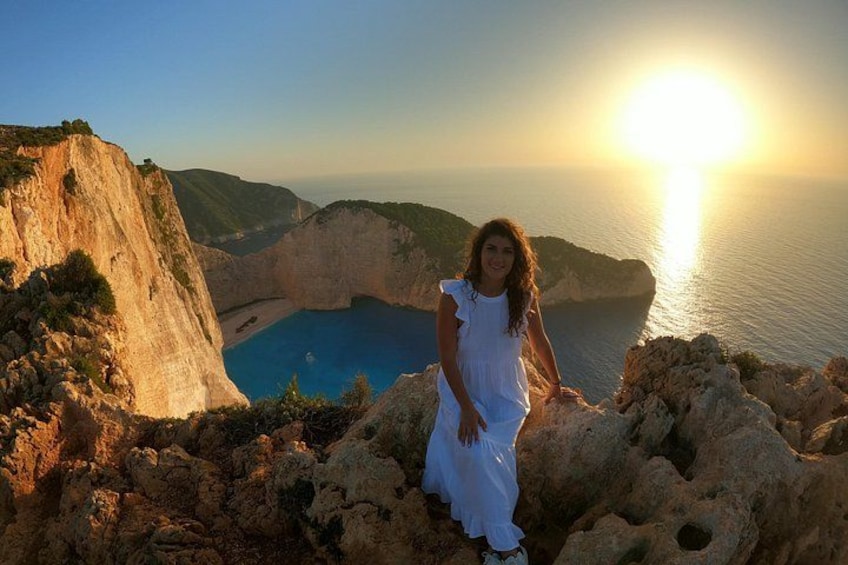 Eine Person in einem weißen Kleid sitzt auf felsigem Gelände, im Hintergrund eine Küstenlandschaft und Klippen. 2