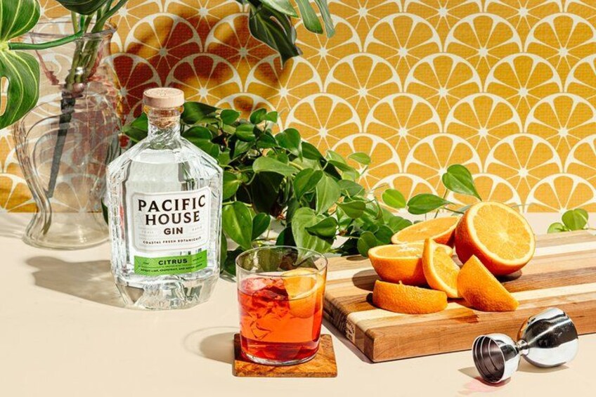 A bottle of Pacific House Gin, a glass of orange juice, and orange slices on a wooden board. 2