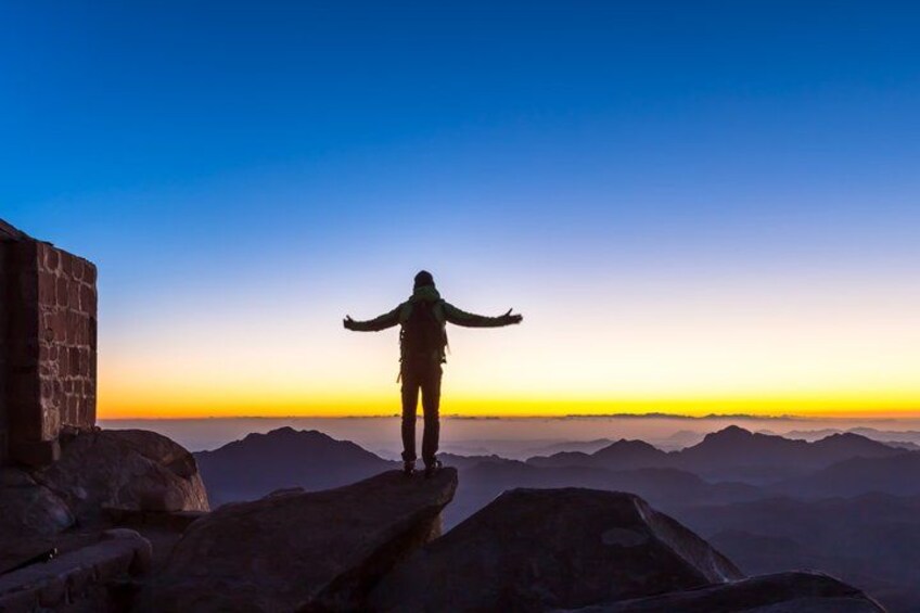 A person standing on a rocky outcrop with arms outstretched, overlooking a mountainous landscape during sunset. 1