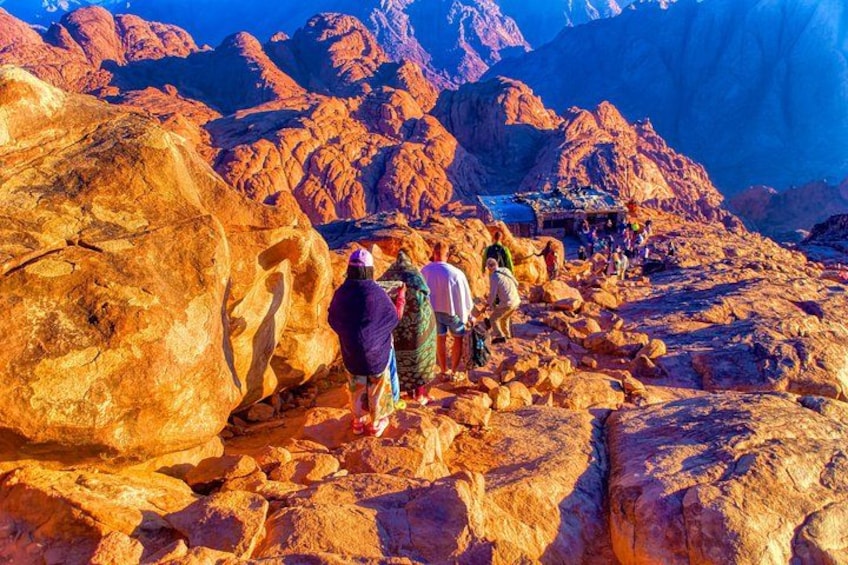 A group of people hiking on rocky terrain with large boulders and mountains in the background. 4