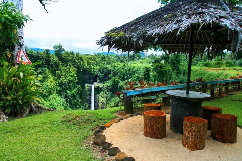 A tropical garden with a thatched-roof gazebo, a waterfall, and a paved walkway. 2