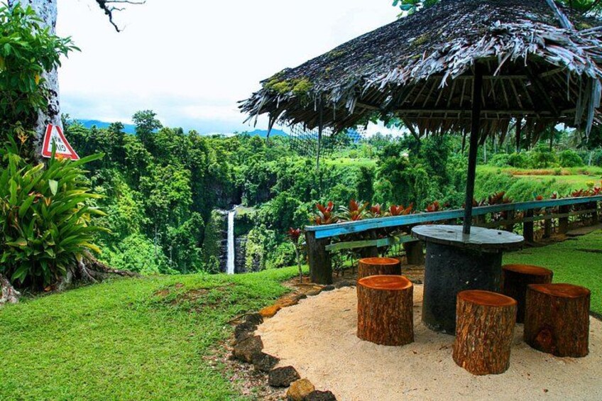 A tropical garden with a thatched-roof pavilion, a waterfall, and a paved walkway. 2