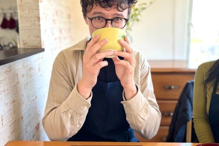 Une personne tenant une tasse jaune, vêtue d’une chemise beige et d’un tablier foncé, est assise à une table dans un café dont les murs sont en briques et le mobilier en bois. 2