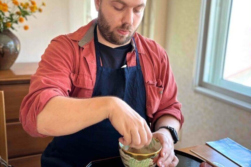A person wearing a red shirt and a dark apron, stirring a green liquid in a bowl. 2